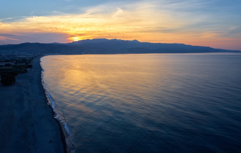 Aerial view from drone on Platanias beachfront area, beach and sea at sunset time. Rodopos peninsula is on background. Chania prefecture, Crete, Greece.
